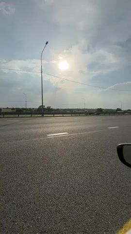 The video is a first-person perspective shot from a moving vehicle on a highway. The sun is visible in the sky, shining through clouds. The road is asphalt with white lane markings. On the right side of the road, there are parked cars, and a yellow line borders the shoulder. A car's side mirror is visible in the foreground, reflecting the road and sky. In the distance, there are streetlights and a line of trees. Traffic appears to be light, with a few cars visible on the road.