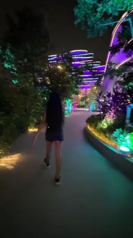 A person with long dark hair and tattoos on their arms walks away from the camera on a paved path at night. They are wearing a dark top and dark shorts. The path is illuminated by colorful lights, including green and purple lights on the surrounding trees and buildings. The buildings in the background have illuminated wavy lines of lights in purple and green. The environment appears to be an outdoor walkway or plaza with lush vegetation and modern architecture.