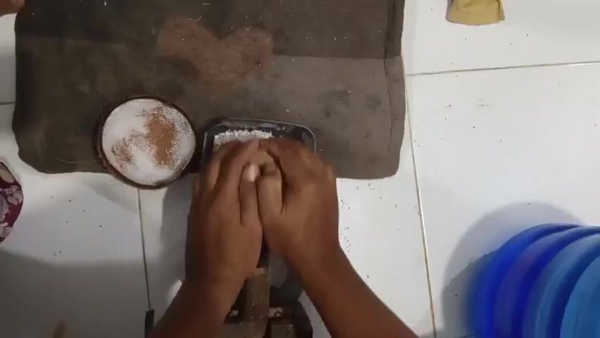 The video shows a close-up, overhead view of a person's hands grating coconut. The person is using a traditional coconut grater, which consists of a wooden base with a metal grating surface. The person is holding a piece of coconut in their hands and rubbing it against the grating surface, producing fine shreds of coconut that fall into a metal container below. To the left of the grater is a half-shell of coconut, also filled with shredded coconut. The grating surface appears to be sharp and is causing the coconut to break down into smaller pieces. The hands are stained brown, suggesting they may have been exposed to coconut husk or other natural materials. The background is somewhat blurred, but it appears to be a tiled floor and a dark brown mat with a heart shape on it. To the right, a blue plastic container is partially visible. The lighting is natural and diffused, indicating the scene is likely outdoors or in a well-lit indoor space. The overall impression is one of traditional food preparation.