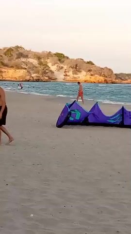 The video shows a beach scene. Initially, there are two women in swimsuits and two people, a man and a child, standing near a blue object. The scene then shifts to the ocean, showing a red ball floating in the water. Later, an older man wearing a light-colored shirt, shorts, and a cap walks barefoot along the shoreline. The beach has light-colored sand, and the ocean is wavy under a pale sky.