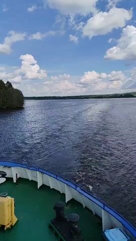 The video shows a view from the back of a boat moving on a wide, dark blue river. The boat has a green deck and a blue and white railing. The water is choppy behind the boat, showing its wake. On either side of the river is land covered in green trees. The sky is blue with white, puffy clouds. The day appears to be sunny.  The camera is stationary on the boat, capturing the scenic view as the boat moves forward.