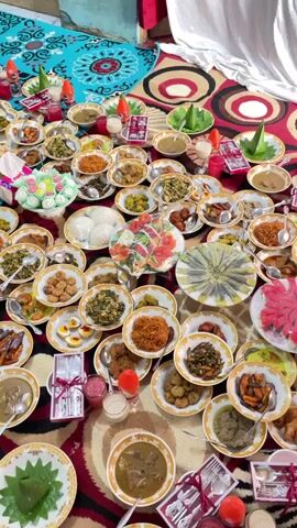 A high-angle shot shows a large spread of individual plates of food arranged on a patterned rug. The plates are diverse, containing various dishes, including what appears to be rice, meats, vegetables, and possibly some desserts. Some plates have cones of green leaves, and others have small bundles of cutlery. There are also several small glasses and bottles scattered among the food. The overall impression is that of a feast or a catered event.