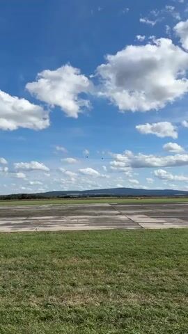 The video shows an outdoor scene with a clear blue sky filled with white clouds. In the foreground, there is a grassy area followed by a tarmac or runway. In the distance, there is a line of trees and hills. Two small aircraft are visible flying in formation against the sky, leaving faint contrails. The camera is stationary and the video captures the aircraft in flight.