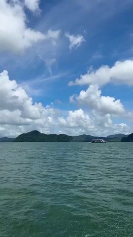 The video shows a wide shot of a body of water under a partly cloudy blue sky. In the distance, several green, mountainous islands are visible. A white boat with a red stripe is moving from left to right across the frame, leaving a slight wake. The water is a deep green and appears slightly choppy. The clouds are white and fluffy, scattered across the sky.