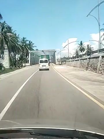 The video shows a view from inside a car driving on an asphalt road, approaching a metal bridge under a clear blue sky. Tall palm trees line the roadside. A white van drives in the same direction ahead of the camera car, on the same side of the road separated by a solid white line. The bridge's structure casts geometric shadows on the road. The bridge has a sturdy metal framework above the road. On the far side of the road, a stone retaining wall is visible beyond the bridge's side rail. There are power poles and street lamps beyond the retaining wall. The dashboard of the camera vehicle is visible in the foreground.