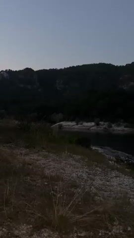 The video shows a dark, natural landscape at dusk or dawn. In the foreground, there is rocky, dry ground with patches of grass. The middle ground features a body of dark water, possibly a lake or reservoir, with a shoreline dotted with white rocks and vegetation. Beyond the water, a dark, tree-covered hill rises, with a lighter sky above it. The overall impression is one of stillness and natural beauty in low light conditions. There are no people or specific actions depicted.