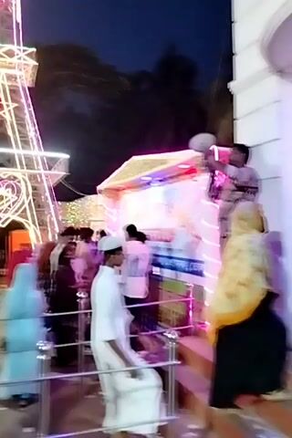 The video captures a lively nighttime scene at what appears to be a public event or fair in Charfasson, Bangladesh.

The central and most prominent object is a large, illuminated replica of the Eiffel Tower, constructed from lights. This structure features a distinct heart shape in its middle section and is adorned with various colored lights, including pink, yellow, and white, creating a festive glow against the dark sky. Its apex also glows brightly.

Numerous people are present in the frame, milling about the area. The crowd comprises adults and children of various ages, suggesting a family-friendly atmosphere. Many individuals are dressed in what appears to be traditional attire; for instance, some men are seen in white panjabis, and some women wear head coverings or burqas.

On the right side of the frame, there's another brightly lit stall or structure, adding to the illuminated environment. Metal railings are visible in the foreground, separating the viewer's perspective from the main crowd.

The activity mainly involves people walking, standing, and observing the illuminated attractions. The overall mood appears to be celebratory and social, with individuals enjoying the festive ambiance created by the lights. The event is clearly taking place after sunset, as evidenced by the dark sky and the strong reliance on artificial lighting. No discernible text or specific location markers other than the Eiffel Tower replica are visible within the video itself.