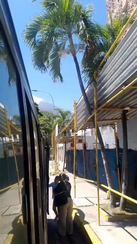 The video shows a side view from inside a vehicle, looking out at a narrow path next to the vehicle. There are people on the path. The path is lined with scaffolding covered with corrugated metal. Palm trees are visible overhead. A mountain is in the background. The people appear to be walking along the path and interacting with each other. One person is reaching for the vehicle door. Another person is walking away from the vehicle. The sky is blue and the sun is shining.