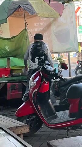 A young man wearing a light-colored long-sleeved shirt and dark pants stands next to a red motorcycle parked under a makeshift awning. He is holding a white plastic bag in his right hand and appears to be interacting with a vendor off-camera to his left. A black helmet rests on the handlebars of the motorcycle. In the background, other motorcycles are visible, along with a street scene featuring buildings and a sign with Indonesian text. The lighting suggests it is daytime.