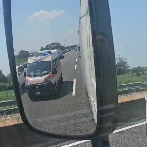 The video appears to be filmed from inside a vehicle, looking into a side mirror. The mirror reflects an ambulance driving on a highway. The ambulance is white with orange and red accents, and has blue flashing lights on top. It is labeled "AMBULANZA." Other vehicles, including cars and a truck, are also visible on the highway. The highway is paved and has white lane markings. The surroundings appear to be rural, with fields and trees visible in the distance. The sky is blue with some clouds. The video quality is somewhat grainy, and there is a slight tilt to the camera.