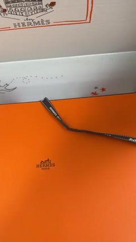 A hand wearing a white sweater opens a box. The box has an orange exterior and a white interior. The inside of the lid has a drawing of a building and the words "chez HERMÈS". Inside the box are two fabric bags with the Hermès logo and white tissue paper. The hand reaches into the box and touches one of the fabric bags.