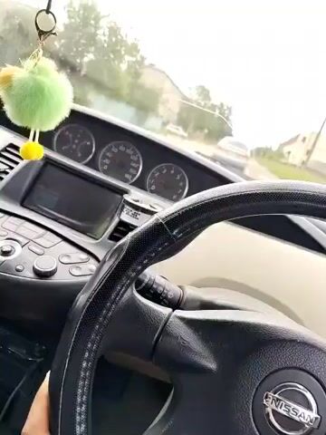 The video is taken from the perspective of someone inside a car, looking at the dashboard and steering wheel. A fuzzy green keychain with a yellow pom-pom hangs from the rearview mirror. The car is in motion, as evidenced by the blurred background and the movement of other vehicles. The driver's hand is visible on the steering wheel. The dashboard shows the speedometer and other gauges. The interior of the car appears to be a Nissan. The environment outside the car suggests an urban or suburban setting with buildings and trees visible.