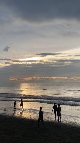 The video shows a beach scene at sunset with several children playing soccer. The sun is low in the sky, casting a warm glow on the water and the sand. The clouds are dark and dramatic, with hints of orange and pink. The children are silhouetted against the bright sky, their movements fluid and energetic as they chase the ball. The waves are gently lapping at the shore, creating a peaceful and serene atmosphere. The overall mood of the video is one of joy and freedom, with the children lost in their game and the beauty of nature.