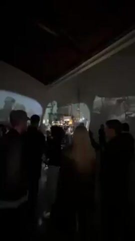 Here is a description of the video:
The video shows a dimly lit room filled with people. Images are projected onto the walls of the room, creating a dynamic and immersive environment. People are standing around, some are silhouetted against the projections, and some moving through the space. The projections appear to be black and white film clips, interspersed with colored scenes. In the center of the room there seems to be a drum set. The room appears to be a gallery space.