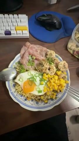The video shows a plate of food on a wooden table. The plate contains instant noodles, a fried egg, corn, shrimp, and ham. A fork is being used to eat the food. In the background, there is a keyboard, a computer mouse, and a jar of food. The lighting is dim.