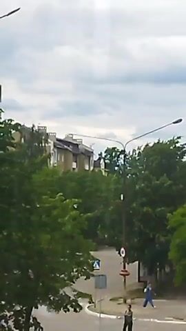 This is a video of a street scene. In the foreground, there are trees and a street lamp. In the background, there are buildings and a cloudy sky. There is a person walking in the distance. The video is shaky, suggesting it may have been filmed handheld.