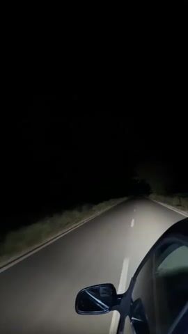 A first-person perspective video shows a car driving down a dark, empty road at night. The headlights illuminate the asphalt road ahead and the sparse vegetation on the sides. The car's side mirror is visible in the lower right corner of the frame. The sky is completely black, indicating a moonless night or heavy cloud cover. There are no other vehicles or people visible in the frame. The camera appears to be mounted inside the car, looking forward and slightly to the right.