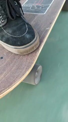 The video shows a close-up view of someone riding a skateboard. The person's left foot, clad in a black suede shoe with black laces and a tan sole, is positioned on the deck of the skateboard. The skateboard deck is brown with a sticker towards the tail end. The wheels are light gray and are spinning as the skateboard moves across a smooth, multicolored surface. The surface changes from green to white and then to red. Shadows move across the ground as the skateboard rolls.