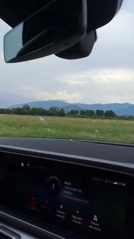 This media file captures a point-of-view perspective from inside a car, looking out through the windshield. The scene is dominated by a wide expanse of green farmland stretching towards a line of trees and distant, hazy mountains under a cloudy sky. The time of day appears to be late afternoon, given the quality of the light and the display on the car's infotainment system showing "17:49". The weather is overcast, suggesting a potentially humid or rainy day.

The car's interior is visible in the foreground, with a prominent rearview mirror at the top and a dashboard featuring a digital display. The display shows information related to a call or contact, with text such as "Juby It", a phone number "+393342089103", and "Telefono". There are also icons for "Numerico", "Aggiorna chiamata", and "Passare alle vettura" suggesting options related to dialing, call management, and vehicle settings.

The road ahead curves to the right, bordered by the farmland on the left and the road on the right. A triangular road sign with a red border and a symbol inside is visible on the right side of the road, indicating a potential hazard or restriction. The overall impression is one of a scenic drive through the Italian countryside, likely near Pinerolo. There are no people visible, only the interior of the car and the external landscape.