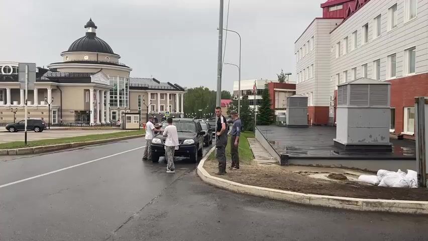 A group of four men stand by two parked cars on a wet asphalt road. One man on a bicycle with a guitar case on his back passes by the group. In the background, there is a large building with a dome and columns, a modern brick building, and some trees. The sky is overcast.