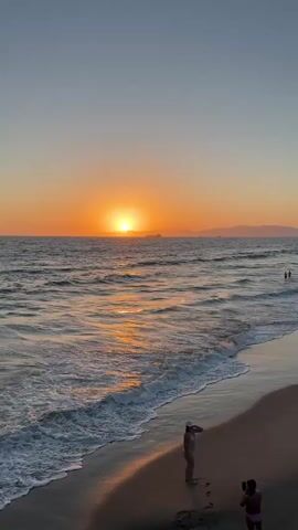A vertical video shows a serene beach scene at sunset. The sun is a bright orange orb on the horizon, casting a golden glow across the rippling ocean waves. The sky above transitions from a pale blue to a soft orange near the horizon. On the sandy shore, two individuals are visible. One person, in the foreground, is facing away from the camera, standing near the water's edge. Another person is further back, also facing away, appearing to hold a phone, possibly taking a photo. A few distant figures can be seen wading in the water. The gentle waves crash onto the wet sand, creating white foam. The overall atmosphere is peaceful and tranquil.