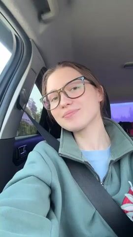 A young woman wearing glasses and a green jacket is in the driver's seat of a car. She's looking directly at the camera and smiling slightly. The car is in motion, as evidenced by the blurry scenery outside the window. The woman's seatbelt is fastened. There is music playing in the background. The interior of the car appears typical, with a dashboard visible and a hint of purple light near the rear window.