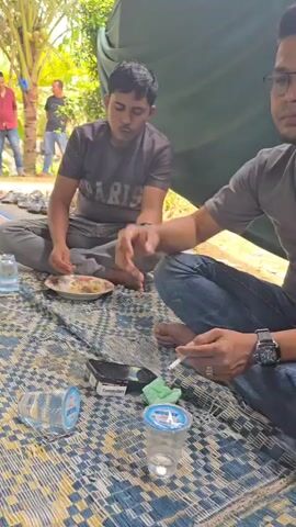 This video captures a gathering of people seated on mats, likely outdoors under a tent or canopy. In the foreground, a young man in a dark t-shirt and black pants is seated cross-legged, holding a plate of food and looking to his right. To his left are two plastic cups, one containing a clear liquid, and a crumpled green tissue. Behind him, other individuals are visible, some wearing headscarves. In the middle ground, a man in a grey shirt and jeans is seated, holding a cigarette. Plates with leftover food and cutlery are scattered around him, along with a pack of cigarettes and a small box. Further in the background, more people are seated, and beyond them, trees and foliage are visible. Two men stand in the far background near some palm trees. The overall impression is of a casual social gathering, possibly a meal or a break.