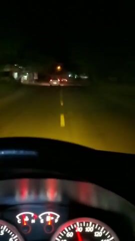 The video is a first-person view from inside a car driving at night. The headlights of the car illuminate a two-lane road with yellow dashed lines marking the center. Several other cars are visible ahead, their taillights glowing red. The dashboard of the car shows red illuminated gauges. The surroundings are dark, with occasional lights from buildings or streetlights visible on the sides of the road. The yellow tint of the headlights suggests they are either fog lights or have a yellow bulb. The car appears to be moving at a moderate speed.