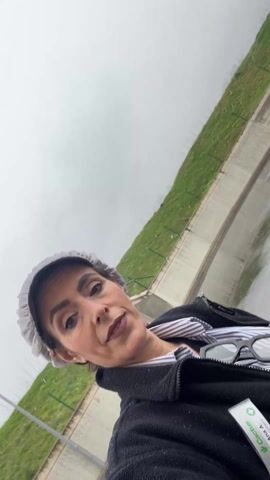A woman in a gray disposable hat and a black jacket stands outside. She is looking directly at the camera and speaking. In the background, there is a low concrete wall topped with a metal railing. Beyond that is a green hillside. The sky is overcast, and it appears to be raining or snowing lightly. She has a pair of glasses tucked into her jacket. The woman is holding a package with the word "Cactus" visible.
