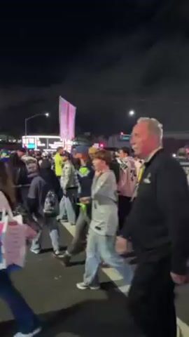 The video shows a crowd of people at night, likely at an outdoor event. Many are wearing Seattle Seahawks jerseys or gear, indicating a sports-related gathering. There are also people holding shopping bags with bright graphics, possibly merchandise. A person holding a sign is visible in the background. The environment appears to be a city street, with buildings and traffic lights visible. A rhythmic noise can be heard in the background. The people are moving and interacting with each other.