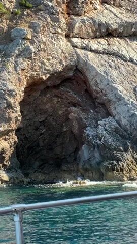A boat moves past a rocky cliff face with a cave opening. The water is a vibrant turquoise and waves gently lap at the base of the cliff. The cliff is rough and textured, with layers of rock visible. Sunlight illuminates the scene, creating shadows and highlights on the rock.