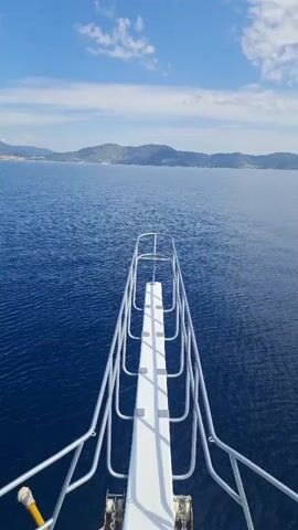 The video shows a forward-facing view from the bow of a boat moving across a large body of water under a clear blue sky with scattered white clouds. In the foreground, the white bow of the boat is visible, featuring a central white plank-like structure and a surrounding metal railing system. The water is a deep blue color, showing gentle ripples as the boat progresses. In the distance, a long stretch of land with rolling hills or mountains is visible under the horizon, suggesting a coastline or large island. No people or visible text are present in the frame. The scene conveys a sense of calm and openness, typical of a journey at sea on a bright day.