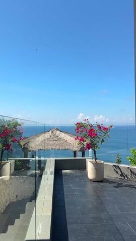 The camera pans right to show a tiled balcony area with a thatched-roof gazebo in the background and the ocean in the distance. Two potted bougainvillea plants flank the gazebo, with one on either side. A glass railing lines the left side of the balcony, while a stone wall is on the right. The sky is clear and blue with a few scattered clouds.