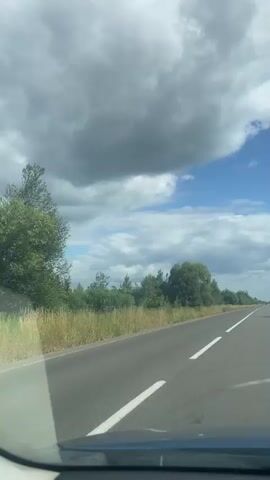 The video shows a first-person perspective of driving on a straight, paved road with a white line marking the lane. The road is flanked by grassy verges and dense green trees. The sky above is partly cloudy, with large, dark gray clouds interspersed with patches of bright blue sky. Sunlight filters through the clouds, casting dappled shadows on the road. The overall impression is of a journey through a rural or semi-rural landscape on a day with mixed weather.