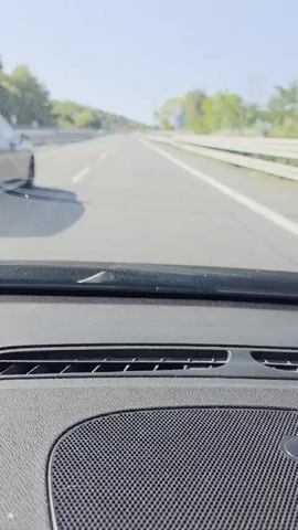 The video displays a forward-facing view from inside a moving vehicle on a multi-lane highway under a clear blue sky. The top of the car's dashboard is visible in the foreground. Ahead, a dark-colored SUV is driving in the left lane, and a lighter-colored station wagon or SUV is in the right lane. The dark SUV overtakes the lighter vehicle. White lane markings delineate the lanes, and a white guardrail runs along the right side of the highway. Trees and hills are visible in the distance.