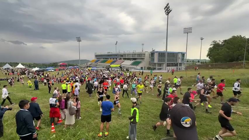 The video shows a large group of people participating in an outdoor event, likely a race, on a grassy field in front of a stadium under a cloudy sky. Many individuals are actively running, while others appear to be in the process of starting or are observing. The participants are dressed in athletic wear, with some wearing brightly colored clothing. There are tents and flags visible in the background, suggesting a race or event setup. The stadium in the background has multiple levels of seating and prominent lighting towers. The ground is covered in green grass, and there are some orange traffic cones on the field. The camera angle is from a slightly elevated position, looking over the crowd towards the stadium.