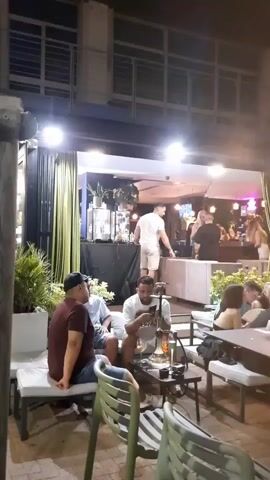 This video shows an outdoor seating area of a bar or restaurant at night. Several people are seated at tables, some of whom are smoking shisha. In the background, a bar area is visible with more people and bar stools. The lighting is dim and ambient, with some spotlights illuminating the bar. A DJ booth is also visible. The overall atmosphere appears to be relaxed and social.