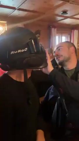 A man is adjusting a black Ruroc ski helmet on another person's head. The person wearing the helmet is facing away from the camera, dressed in a black long-sleeved shirt. The man adjusting the helmet is standing behind them, wearing a dark jacket. They are in a room with a wooden ceiling and hanging light fixtures. In the background, people are seated at tables. There are windows behind the tables. The general atmosphere appears to be casual and indoors.