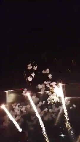 The video shows a nighttime fireworks display over water. Several fireworks are visible, some exploding in bursts of white light and smoke, others forming trails of sparks. The reflections of the fireworks can be seen on the water's surface. A small portion of a person's head and shoulder is visible in the lower left corner of the frame. The overall scene is dark, with the fireworks providing the primary illumination.