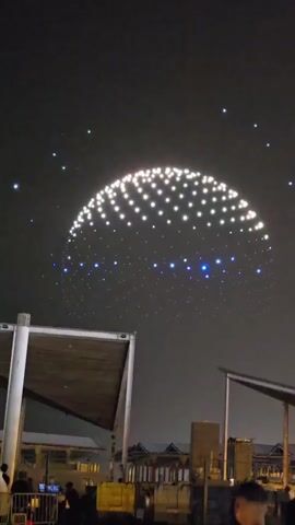 The video captures a nighttime drone light show in a semi-circular formation against a dark sky. Numerous white and blue lights, likely drones, are arranged in a large, glowing dome shape with smaller, scattered blue lights below. In the foreground, two angled awnings are visible, supported by large white poles. Below the awnings, silhouetted figures of people are seen gathering, some looking up at the sky. The background shows a dimly lit urban or waterfront area with buildings and structures. The overall atmosphere is one of a public event or celebration.