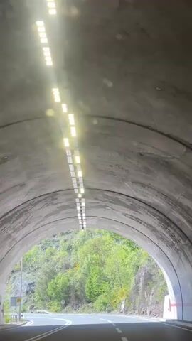 The video shows a car exiting a tunnel on a bright, sunny day. The tunnel is lit with a row of bright, square lights. Outside the tunnel, a road winds along a hillside covered with green trees and rocky outcroppings. The sky is blue with scattered white clouds. Road signs are visible along the roadside.
