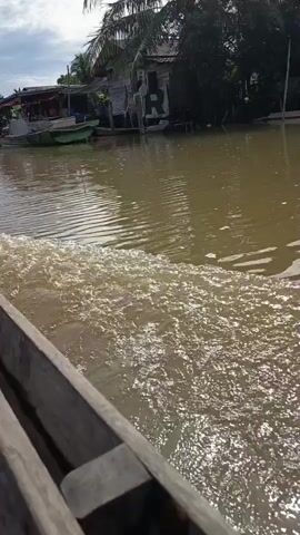 The video shows a view from a wooden boat moving through a muddy river. The camera is angled towards the side of the boat, capturing the churning wake behind it. In the background, there are several other boats docked along the riverbank, with houses and lush green vegetation visible beyond them. The sky is partly cloudy. The scene appears to be a rural riverine community.