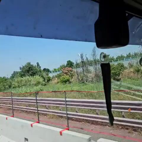 The video is taken from inside a vehicle, possibly a truck or bus, as it travels along a highway on a clear, sunny day. The camera is positioned on the left side of the vehicle, offering views through the windshield and side mirror.

Through the windshield, the viewer sees a multi-lane highway with other vehicles including cars and trucks. A green overhead sign with white lettering spans the highway. Beside the road is greenery with a barrier of white concrete and orange fencing.

The side mirror reflects the same highway and a truck traveling in the same direction. The mirror is visibly dirty with splatters. The overall environment suggests a road trip on a well-maintained highway during daylight hours.