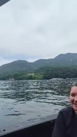 The video shows a group of people on a boat trip. The first frame features a woman smiling and looking to the left. The second frame shows a person wearing a hat and sunglasses, holding a baby in a blue hat. The boat is moving, creating a wake in the water. In the background, there are mountains covered with trees and some buildings along the shore. The sky is overcast. The third and fourth frames continue to show the boat moving through the water with the same background scenery. The camera is mounted on the boat, giving a first-person perspective of the journey.