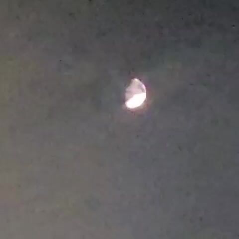 The video opens with a blurry image of the moon in a gray sky. This transitions to a nighttime scene showing a building with lights and a vehicle. The final frames display a hand holding a small, white, crumpled object over a gray, speckled surface.