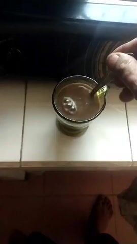 A person's hand is stirring a beverage in a clear glass mug with a metal spoon. The mug is placed on a tiled surface, and a black stovetop is visible in the background. The beverage appears to be light brown in color and is frothy where it is being stirred. The person's bare foot is also visible at the bottom of the frame. The video is a close-up shot, and the lighting suggests it was taken indoors during the daytime. The action of stirring is the primary focus of the video.