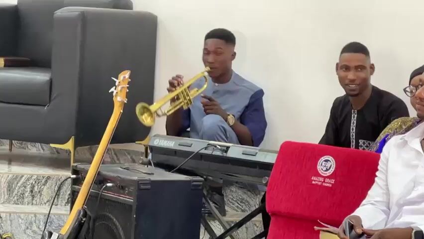 A man in a light blue and dark blue shirt plays a trumpet. He is facing to the right of the frame, and he is sitting in front of a keyboard. To his right, another man in a dark blue shirt and a woman in a black head scarf sit in chairs. To the left of the frame, a bass guitar rests on an amplifier. The background is a white wall and a black couch.