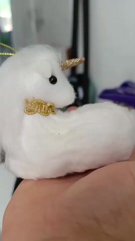 A close-up shot of a person's hand holding a small, white, fuzzy unicorn ornament. The unicorn has a gold glittery horn, black eyes, and a gold collar. The background is blurred and appears to be indoors.