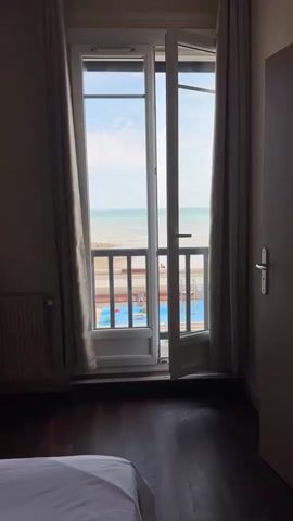 A full shot from a bedroom shows a wide open balcony door with glass panels, revealing a view of the sea and a beach below. The curtains on the left and right sides of the door are drawn back. A white radiator is visible on the left wall, and a dark wooden floor is visible in the foreground. The balcony railing is made of wood, and below it, there is a blue swimming pool with children playing in it. The sky is blue with some clouds. A white bed is partially visible in the bottom left corner.