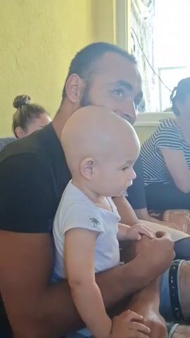 A man with a beard and a shaved head holds a baby on his lap. The man is wearing a black t-shirt. The baby is wearing a white onesie with a palm tree graphic. The baby is looking to the right. The man is looking to the right and smiling. In the background, a woman with dark hair is visible. The environment appears to be indoors, with a yellow wall and a window in the background. The scene is illuminated by natural light.
